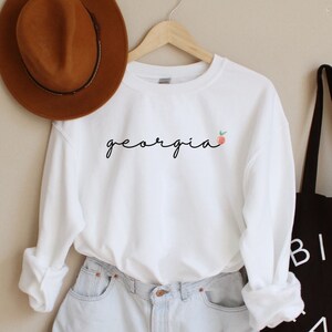May include: White long-sleeved sweatshirt with the word "georgia" written in cursive with a small peach graphic. The sweatshirt is hanging on a hanger and is paired with light blue denim shorts and a black tote bag with the word "BIEN FAIT" printed on it.