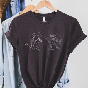 May include: A dark gray t-shirt with a white graphic of a cowboy riding a horse being chased by a dinosaur.