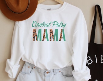 May include: White long-sleeve sweatshirt with the text "Cerebral Palsy MAMA" in green and orange leopard print. The sweatshirt is hanging on a hanger and is styled with denim shorts and a brown hat.