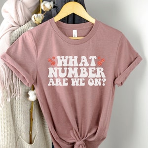 K&ouml;nnte beinhalten: Ein mauvefarbenes T-Shirt mit dem Text "WHAT NUMBER ARE WE ON?" in Wei&szlig; mit roten Herzen auf beiden Seiten des Textes.