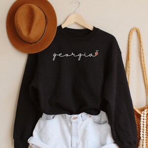 May include: Black sweatshirt with the word "georgia" and a peach graphic in white embroidery. The sweatshirt is paired with light blue denim shorts and a brown woven bag with white beads.