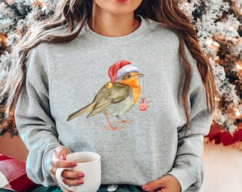 May include: A gray sweatshirt with a watercolor illustration of a robin wearing a Santa hat and holding a red ornament. The robin is perched on a branch with green leaves.