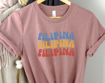 May include: A mauve t-shirt with the word "FILIPINA" repeated three times in a retro font. The word is in blue, yellow, and red.