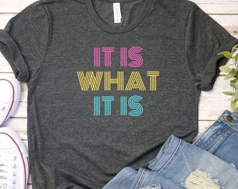 May include: A dark gray t-shirt with the text "IT IS WHAT IT IS" in yellow, pink, and blue stripes. The shirt is laid flat on a white wooden surface with a pair of white sneakers and a pair of blue jeans.