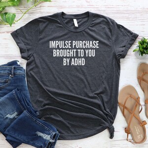 Puede incluir: Una camiseta gris oscuro con el texto "Impulse purchase brought to you by ADHD" impreso en blanco.