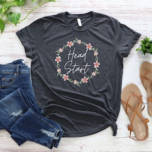 May include: A dark grey t-shirt with a white floral wreath and the text "Head Start" in white.