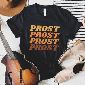 May include: Black t-shirt with the word "PROST" repeated four times in a retro font, with each word in a different shade of orange, yellow, and red. The shirt is laid flat on a white and black striped surface with a guitar, a hat, and a potted cactus.