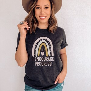 May include: A dark gray t-shirt with a rainbow graphic and the text "I.E.P. I ENCOURAGE PROGRESS".