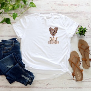 May include: White t-shirt with a brown leopard print heart and the text "EARLY CHILDHOOD" printed on the front. The shirt is paired with blue jeans and brown sandals.