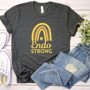 May include: A dark grey t-shirt with a rainbow design and the text "Endo STRONG" in yellow. The shirt is laid out on a white wooden surface with a pair of white sneakers and blue jeans.