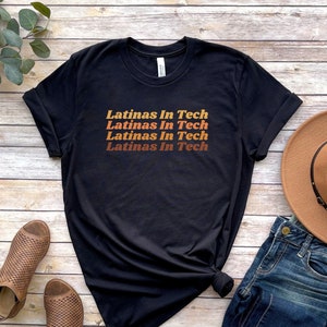 May include: Black t-shirt with the text "Latinas In Tech" repeated four times in a gradient of orange, red, and brown. The shirt is laid flat on a woodgrain surface with a pair of brown sandals, a pair of blue jeans, and a brown hat.