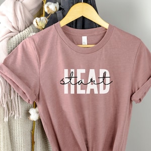 May include: A dusty rose colored t-shirt with the text "HEAD start" printed in white with black cursive writing.