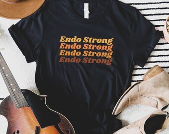 Puede incluir: Camiseta negra con el texto "Endo Strong" repetido cuatro veces en una fuente retro con colores naranja, amarillo y rojo.