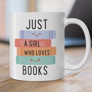 Op de afbeelding: Witte keramische mok met de tekst "JUST A GIRL WHO LOVES BOOKS" en een stapel kleurrijke boeken met glimlachende gezichten.