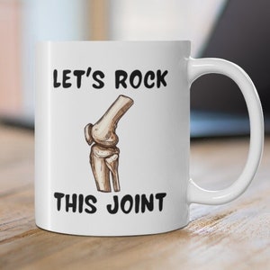 May include: White ceramic coffee mug with a black illustration of a knee bone and the text "Let's Rock This Joint".