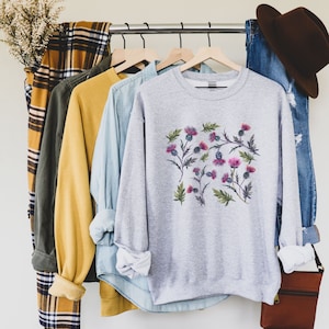 May include: A gray sweatshirt with a floral print of pink and purple thistles. The sweatshirt is hanging on a rack with other clothing.