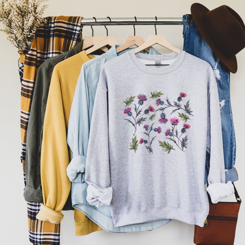 May include: A gray sweatshirt with a floral print of pink and purple thistles. The sweatshirt is hanging on a rack with other clothing.