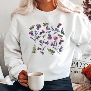 May include: White sweatshirt with a floral print of purple and pink thistles. The sweatshirt has a crew neck and long sleeves.