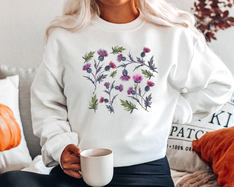 May include: White sweatshirt with a floral print of purple and pink thistles. The sweatshirt has a crew neck and long sleeves.