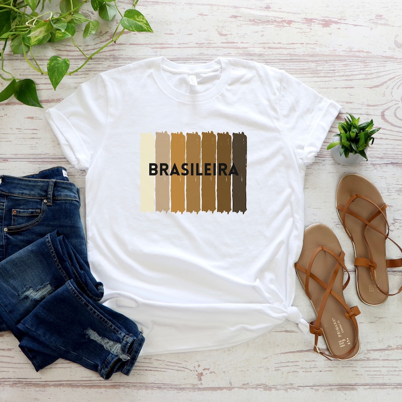 May include: White t-shirt with a graphic of a rainbow of skin tones and the word "BRASILEIRA" in black text.