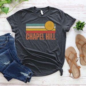 May include: A dark heather grey t-shirt with a retro-style graphic design featuring a sun and stripes in shades of blue, green, yellow, orange, and red. The text "CHAPEL HILL" is printed in white below the graphic.