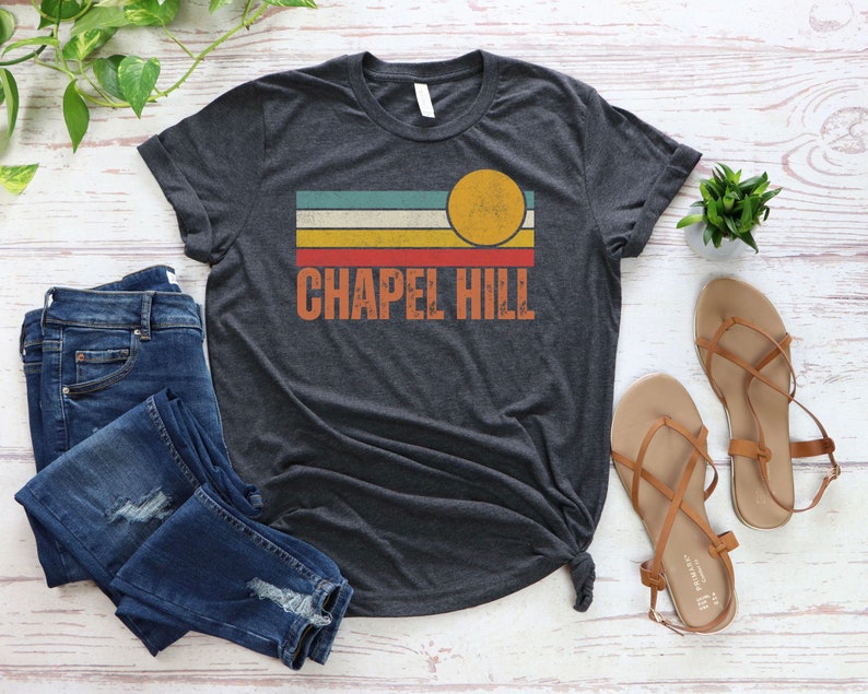 May include: A dark heather grey t-shirt with a retro-style graphic design featuring a sun and stripes in shades of blue, green, yellow, orange, and red. The text "CHAPEL HILL" is printed in white below the graphic.