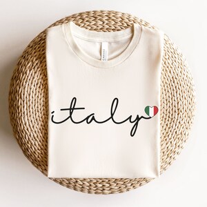 May include: A white t-shirt with the word "italy" written in black cursive script with a small red, white, and green heart with the Italian flag colors next to it.
