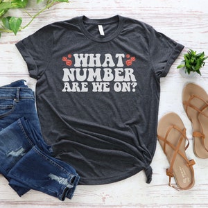 K&ouml;nnte beinhalten: Ein dunkelgraues T-Shirt mit dem Text "WHAT NUMBER ARE WE ON?" in Wei&szlig; mit roten W&uuml;rfelgrafiken auf beiden Seiten des Textes.