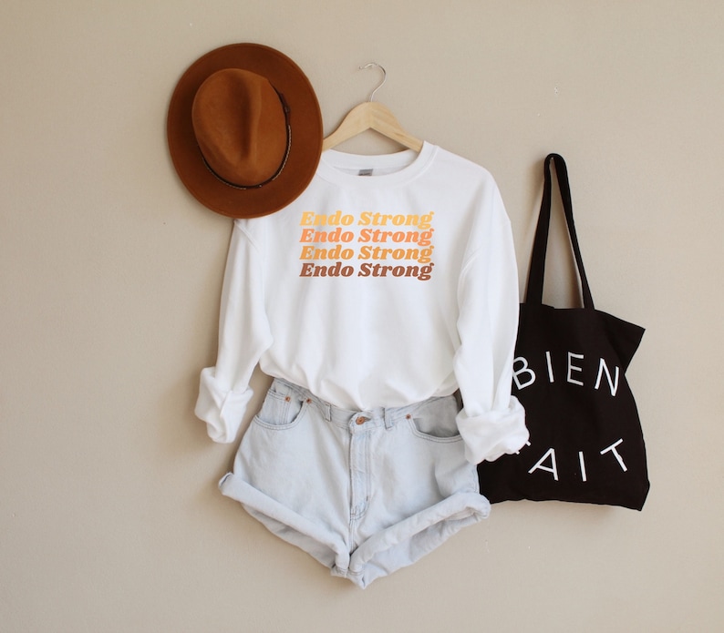 May include: White long-sleeved sweatshirt with the text "Endo Strong" repeated four times in a gradient of orange, yellow, and brown. The sweatshirt is worn with light blue denim shorts and a brown fedora.