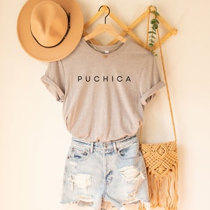 K&ouml;nnte beinhalten: Ein graues T-Shirt mit dem Wort "PUCHICA" in Schwarz auf der Vorderseite. Das T-Shirt h&auml;ngt an einem Holzb&uuml;gel und wird mit Jeans-Shorts und einer Strohtasche kombiniert.
