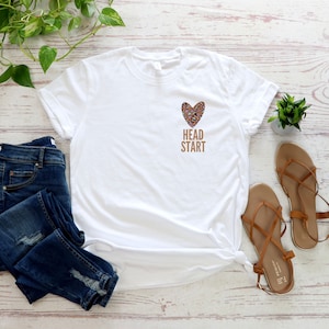 May include: White t-shirt with a brown leopard print heart and the text "HEAD START" in brown lettering.