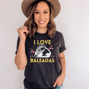 May include: A dark grey t-shirt with the text "I LOVE BALEADAS" in yellow and white. A white illustration of a baleada is in the center of the shirt. There are pink hearts around the illustration.