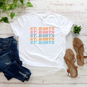 May include: White t-shirt with the text "ST. BARTS" repeated five times in a stacked, retro-style font. The text is in shades of pink, blue, and orange.
