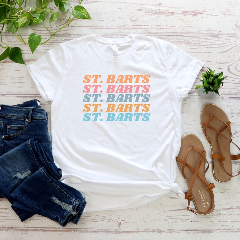 May include: White t-shirt with the text "ST. BARTS" repeated five times in a stacked, retro-style font. The text is in shades of pink, blue, and orange.