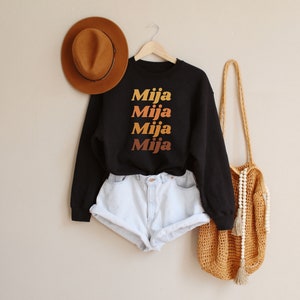May include: Black sweatshirt with the word "Mija" in a gradient of orange and yellow. The sweatshirt is paired with light blue denim shorts. A brown hat and a woven bag with a beaded strap are also in the image.