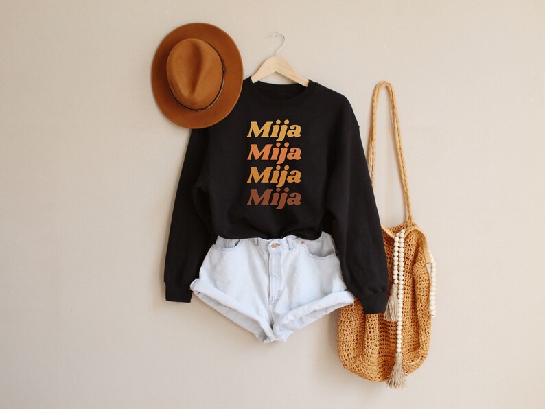 May include: Black sweatshirt with the word "Mija" in a gradient of orange and yellow. The sweatshirt is paired with light blue denim shorts. A brown hat and a woven bag with a beaded strap are also in the image.