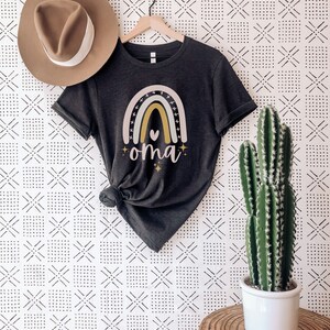 May include: A dark grey heather t-shirt with a rainbow design and the word "oma" in gold lettering. The rainbow has a pink, yellow, and green color scheme with a small heart in the center.