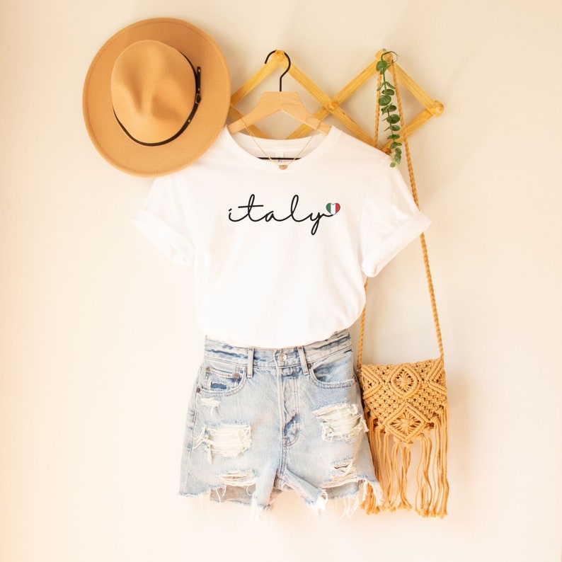 May include: White t-shirt with the word "italy" written in cursive with a small Italian flag heart next to it. The shirt is hanging on a wooden hanger with a straw hat and a woven bag.