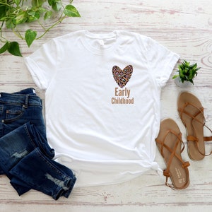 May include: White t-shirt with a brown leopard print heart and the text "Early Childhood" on the front. The shirt is paired with blue jeans and brown sandals.