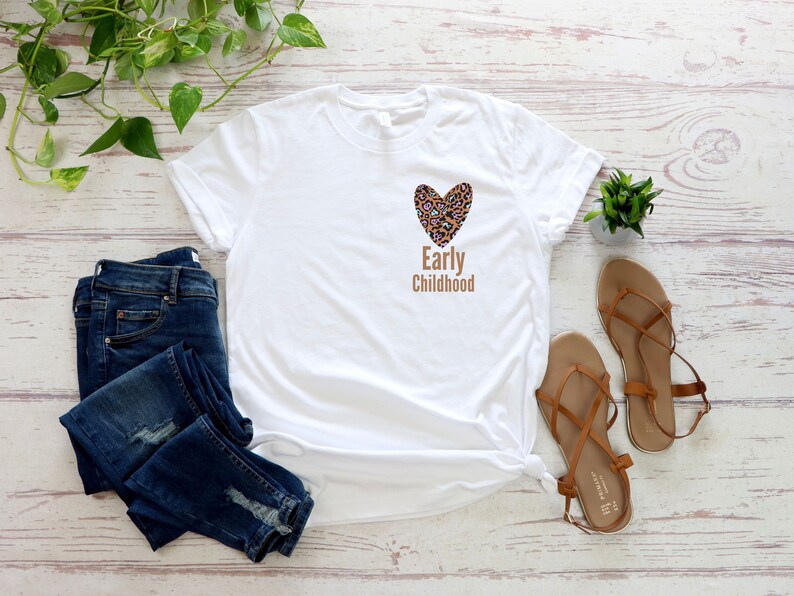May include: White t-shirt with a brown leopard print heart and the text "Early Childhood" on the front. The shirt is paired with blue jeans and brown sandals.
