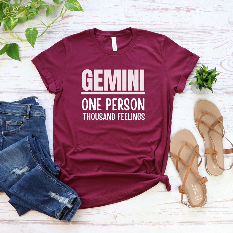 May include: A maroon t-shirt with white text that reads "GEMINI ONE PERSON THOUSAND FEELINGS".