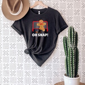 May include: A dark grey heather t-shirt with a gingerbread man graphic wearing a red bow tie and holding a hockey stick. The gingerbread man is in front of a hockey net and the text "OH SNAP!" is printed below the graphic.
