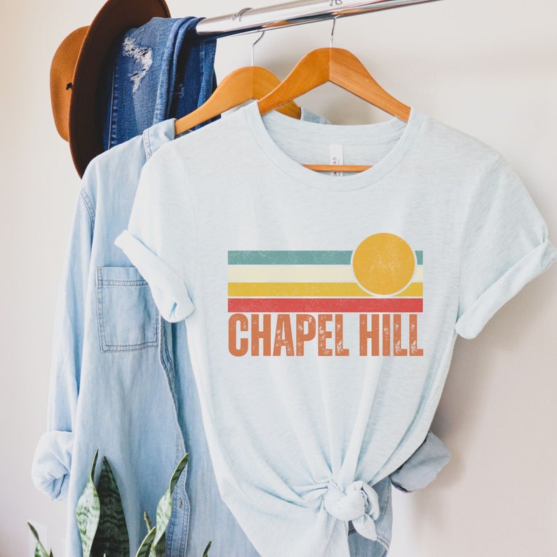 May include: A light blue t-shirt with a retro-style graphic design featuring a yellow sun and colorful stripes. The text "CHAPEL HILL" is printed in orange below the graphic.