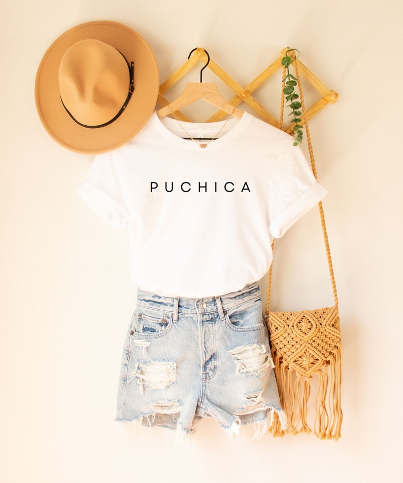 K&ouml;nnte beinhalten: Wei&szlig;es T-Shirt mit dem Wort "PUCHICA" in Schwarz auf der Vorderseite. Das Shirt h&auml;ngt an einem Holzb&uuml;gel mit einem Strohhut und einer gewebten Tasche.