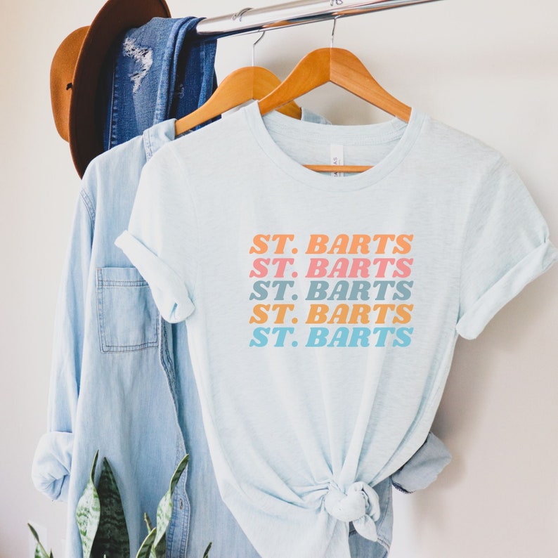 May include: A light blue t-shirt with the text "ST. BARTS" repeated five times in a stacked design. The text is in a retro font and is printed in shades of pink, orange, and blue.
