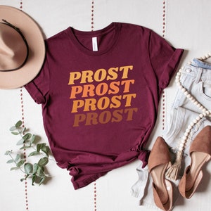 May include: A maroon t-shirt with the word "PROST" repeated four times in a gradient of orange, yellow, and brown. The shirt is laid flat on a white surface with a brown hat, a pair of brown shoes, and a pair of light blue jeans.