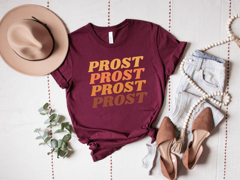 May include: A maroon t-shirt with the word "PROST" repeated four times in a gradient of orange, yellow, and brown. The shirt is laid flat on a white surface with a brown hat, a pair of brown shoes, and a pair of light blue jeans.