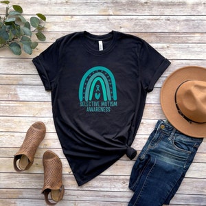 May include: Black t-shirt with a teal rainbow design and the words "Selective Mutism Awareness." The shirt is paired with blue jeans, brown shoes, and a brown hat. The design includes a ribbon and heart.