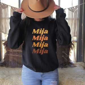 May include: Black crewneck sweatshirt with the word "Mija" in a gradient of orange and brown. The sweatshirt is paired with light blue jeans and a tan fedora hat with a black band.