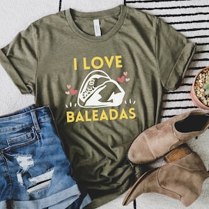 May include: A green t-shirt with the text "I LOVE BALEADAS" in yellow and a white illustration of a baleada. The shirt is on a white and black striped surface with a pair of brown boots and a pair of blue denim shorts.